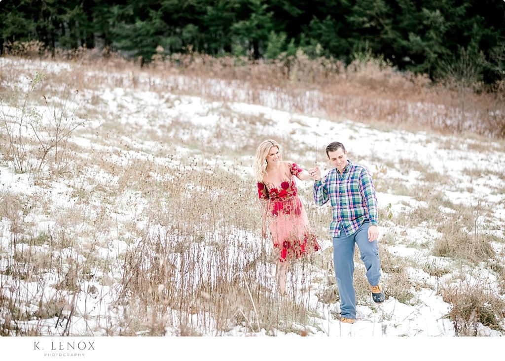 Man helps woman walk down a snowy field during their engagement session by K. Lenox Photography
