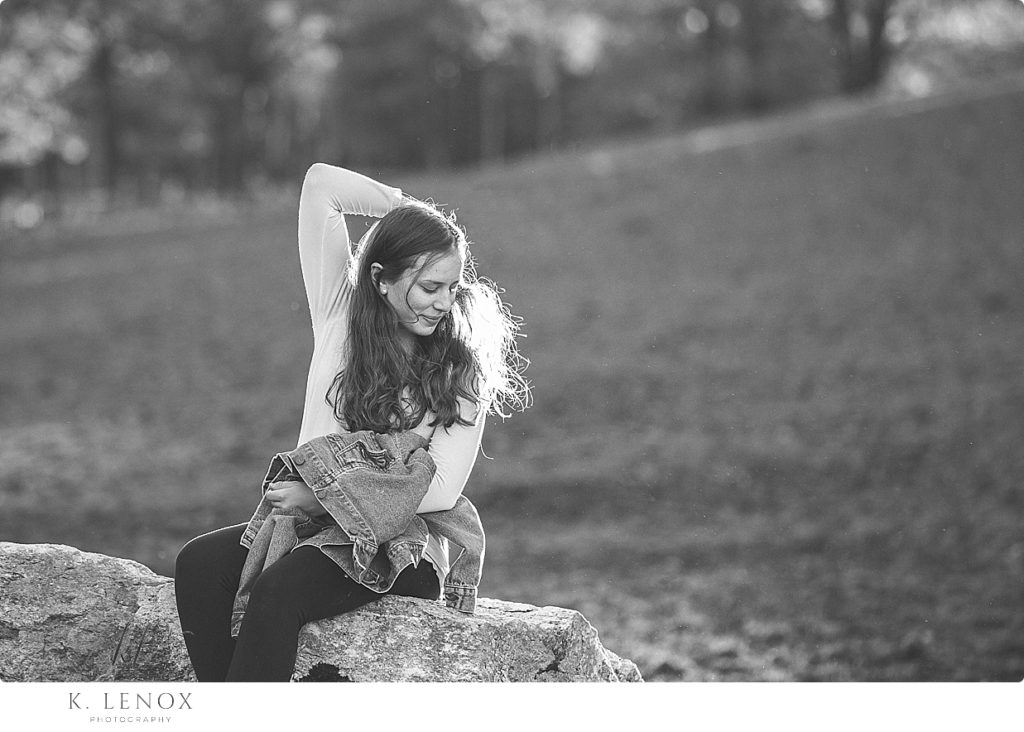 Black and white candid photo of a high school senior girl fixing her hair while sitting on a rock. 