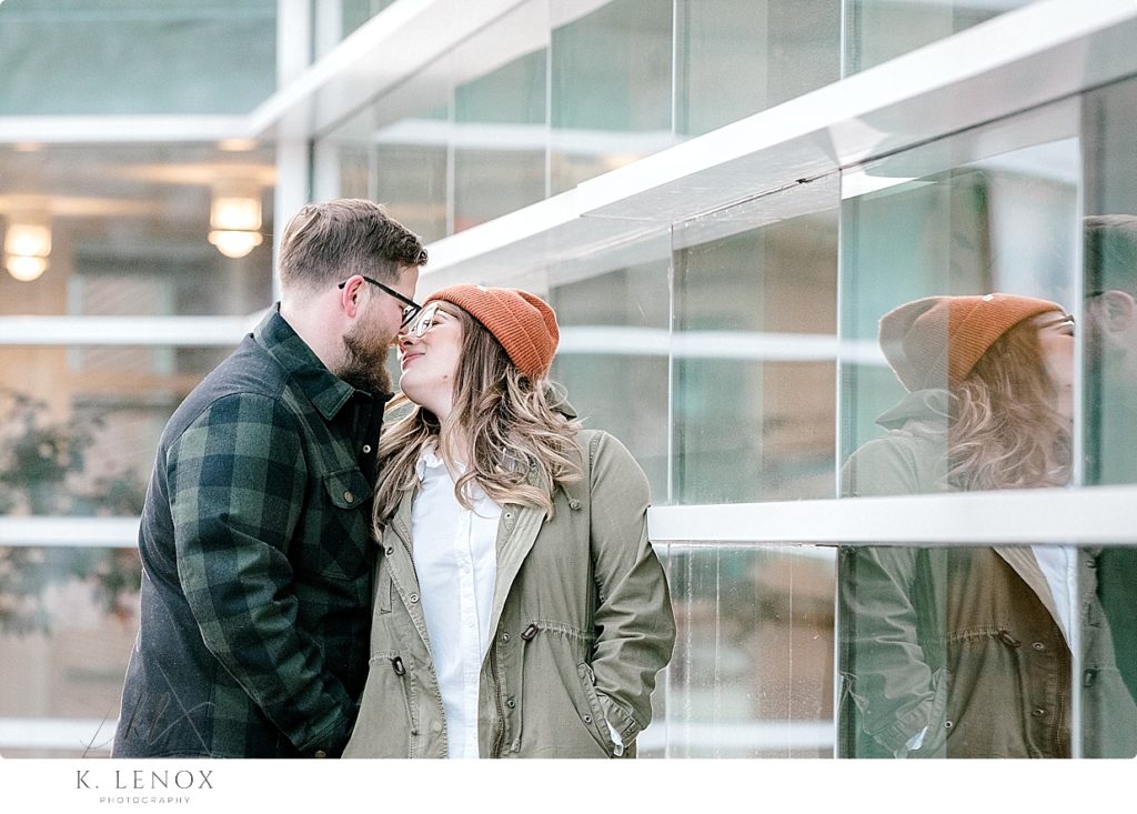 Candid photo of a man and woman kissing near a reflective window for their Light and Airy Winter engagement Session. 