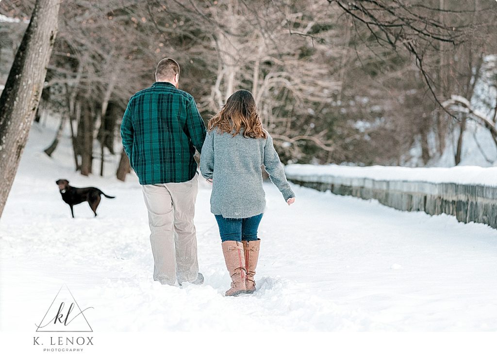 Winter Engagement session with a man and woman walking hand in hand in the snow with their chocolate lab in the distance. 