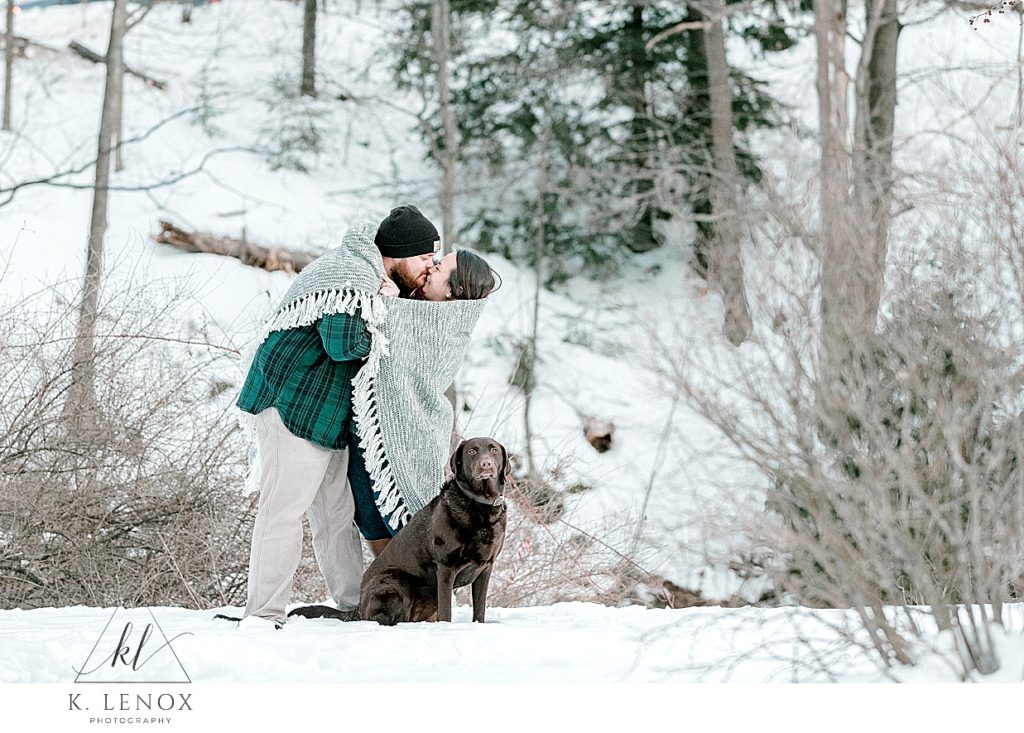 Light and Airy winter engagement session in peterborough.   Couple wrap up in a gray blanket and share a kiss.   Their Chocolate lab sits near their feet. 