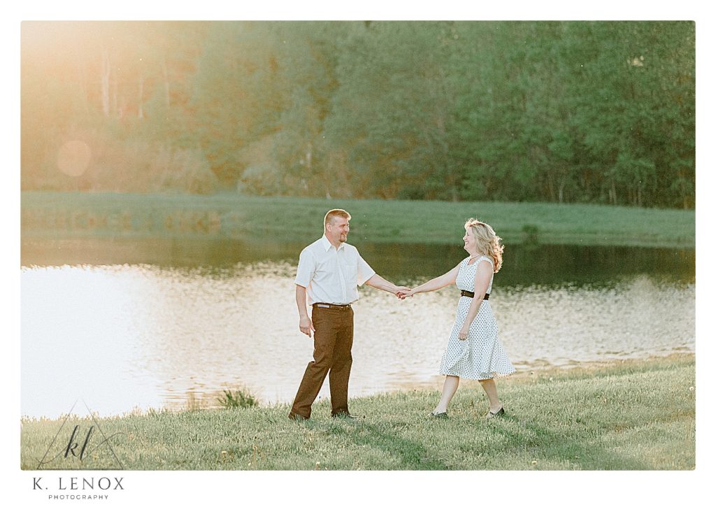 Candid photo of a man and woman (wearing a white dress with black dots)  holding hands in front of a pond with the sun shining behind them.   