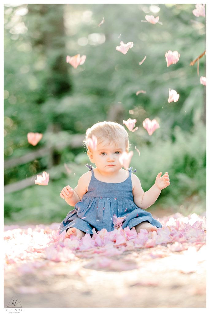 Light and Airy Film inspired photo of a 10 month old little girl wearing a blue dress.  Pink Flower petals are falling around her. 