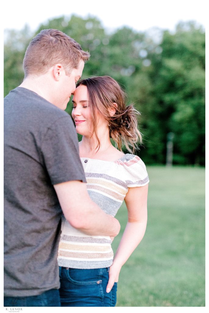Color photo of a casual engaged couple at the park with green trees behind them. 