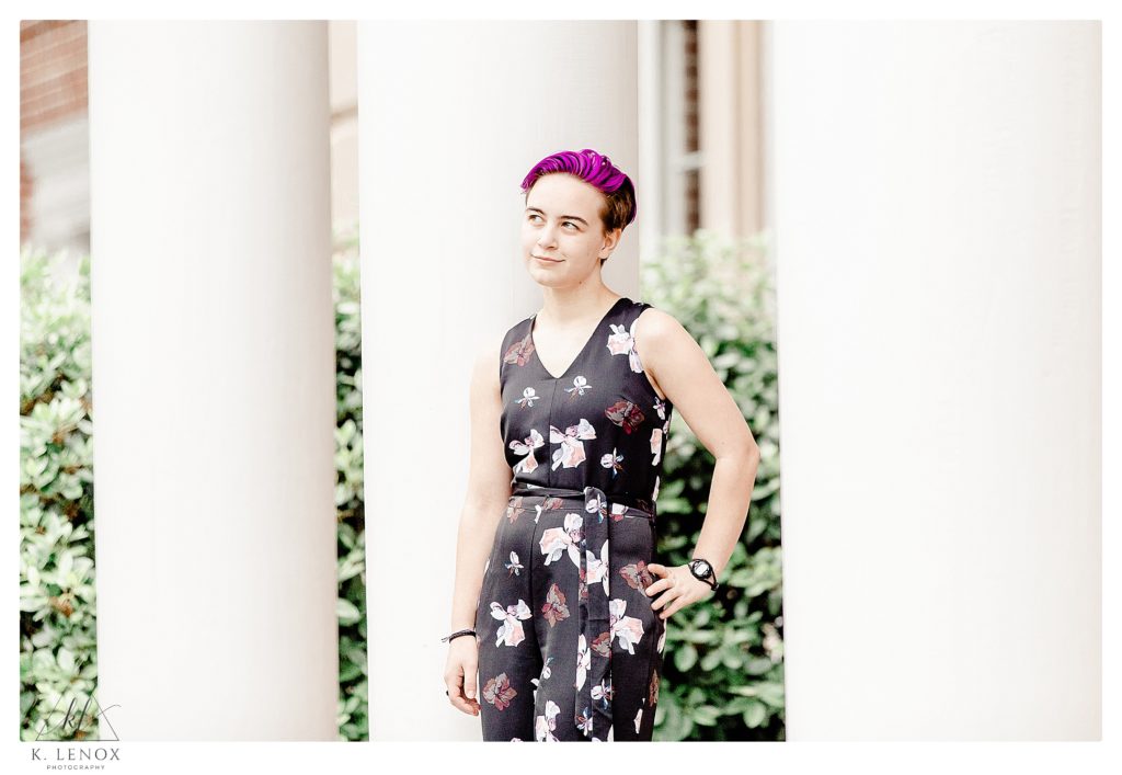 Badass senior portrait of a girl standing in front of white pillars.   Purple hair, and wearing a black floral jumpsuit. 