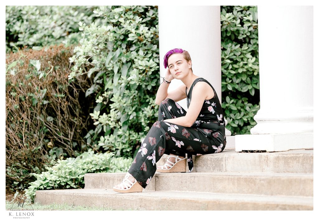 Picture of a girl sitting on concrete steps in front of white pillars.   Purple hair, and wearing a black floral jumpsuit. 