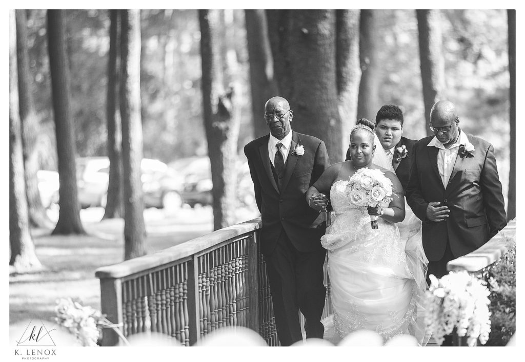 Classic Wedding Ceremony at Alpine Grove in NH.  Black and white photo of a bride walking over the wooden bridge to the ceremony- with her two fathers. 