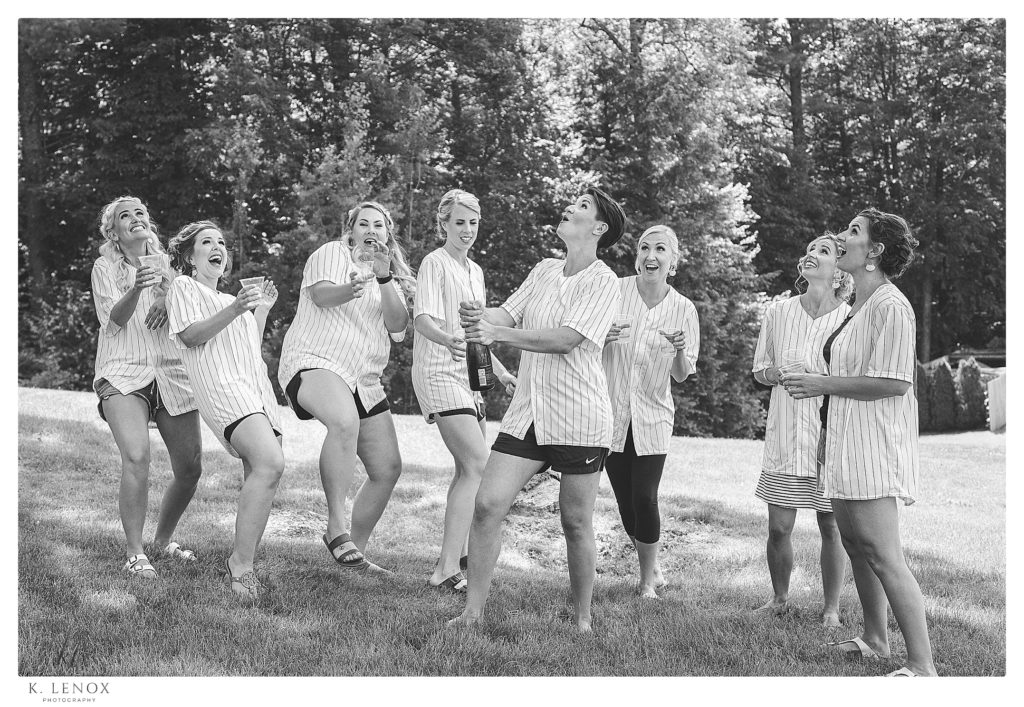 Wedding Party wearing customized Baseball jerseys pops the cork off the champagne and they watch as the cork flies.  Black and white image for a Summer wedding at Mount Sunapee Resort.  