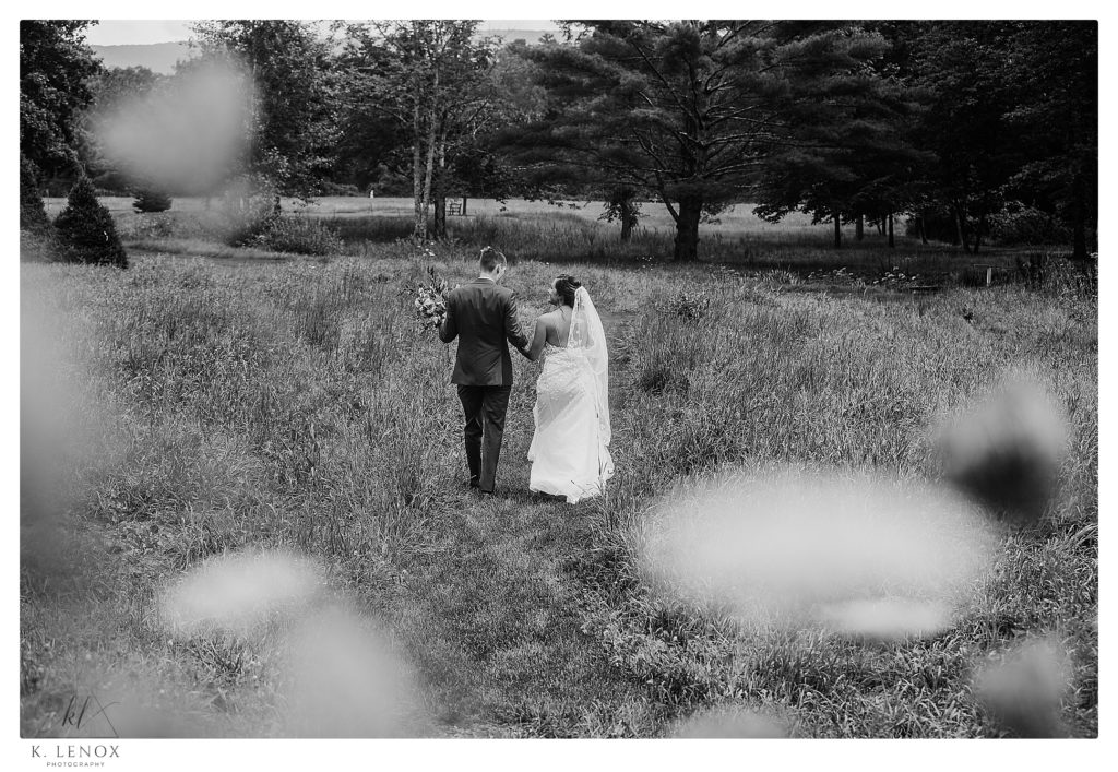 Candid , black and White photo showing a bride and groom walking in a field holding hands. 