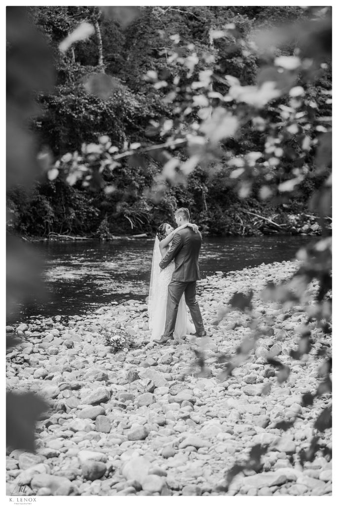 Candid black and white photo showing a bride and groom hug each other next to a river in Vermont. 