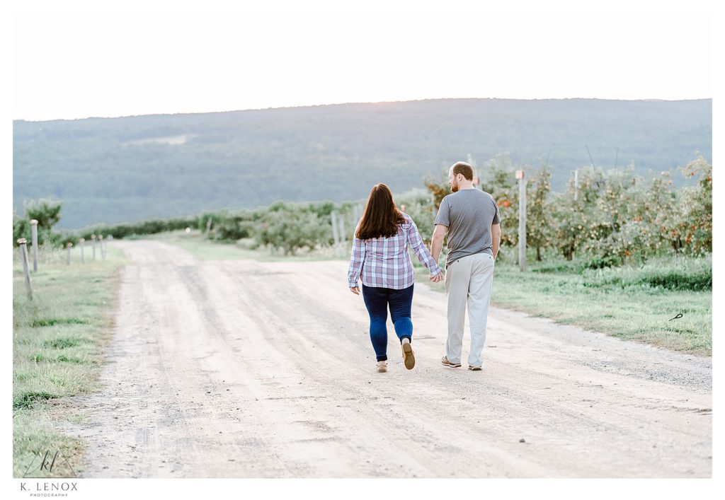Light and Airy Engagement Session at Alyson's Orchard- showing a man and woman walk hand in hand on the dirt road. 