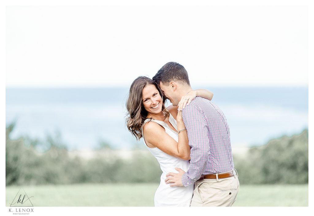 Light and Airy Photo showing a Man and Woman hugging with a view of the ocean in the back ground.  Image taken during an engagement session at the Crane Estates. 