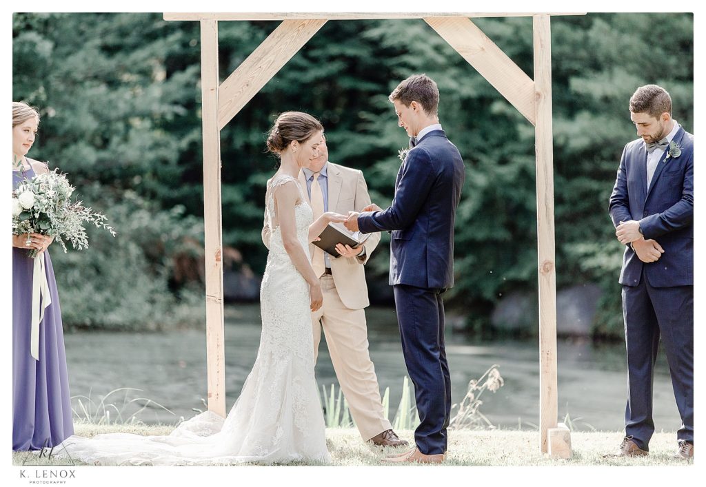 Bride and Groom exchange rings in front of a plain wooden arbor, outdoors for a simple Tamworth NH Wedding