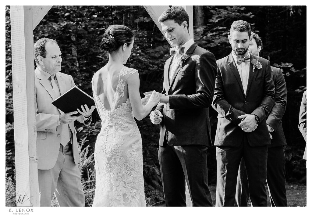 Black and White photo of Bride and Groom exchange rings in front of a plain wooden arbor, outdoors for a simple Tamworth NH Wedding