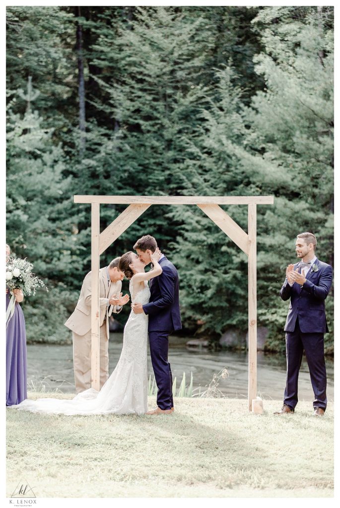 Bride and Groom share their first kiss in front of a plain wooden arbor, outdoors for a simple Tamworth NH Wedding