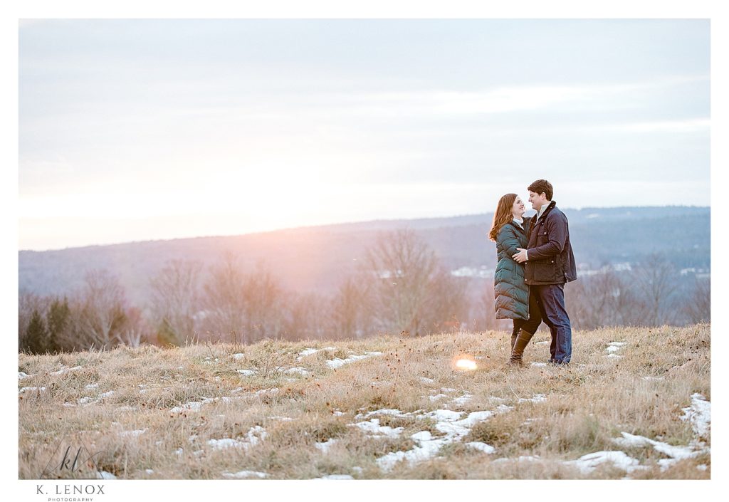 Light and Airy Winter Engagement Session at Alyson's orchard showing a man and woman standing on a hill overlooking the view, as the sun sets. 